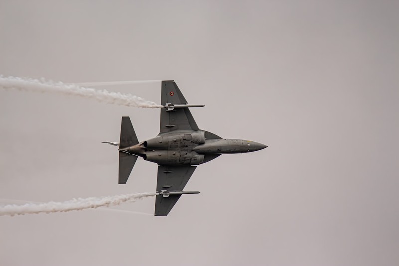 A military fighter jet flies through an overcast sky with white smoke trails streaming from its engines
