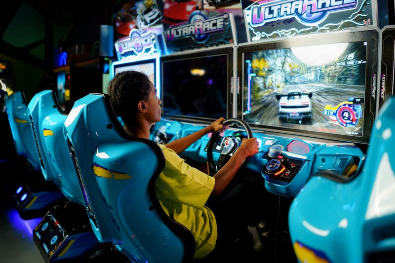 A person playing a racing arcade game, gripping the steering wheel of a blue arcade simulator in a neon-lit arcade