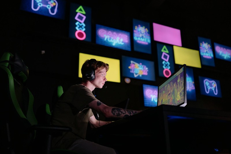 A focused gamer in a neon-lit room playing on a computer with headset and multiple glowing gaming-themed displays