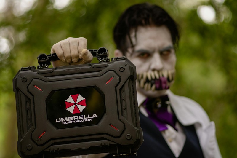 A cosplayer in zombie makeup holding a black case bearing the Umbrella Corporation logo from Resident Evil