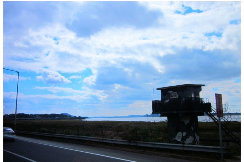 A weathered military observation tower stands near a fence and body of water in the Korean Demilitarized Zone