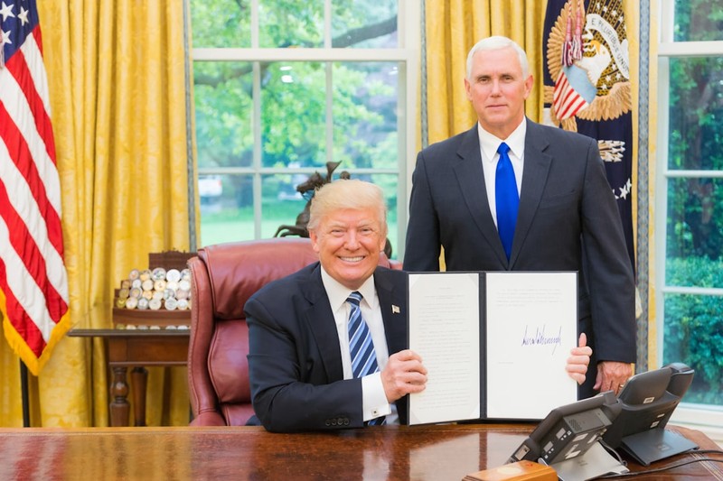 President Donald Trump seated at the Resolute Desk in the Oval Office, signing a document with Vice President Mike Pence standing behind him.