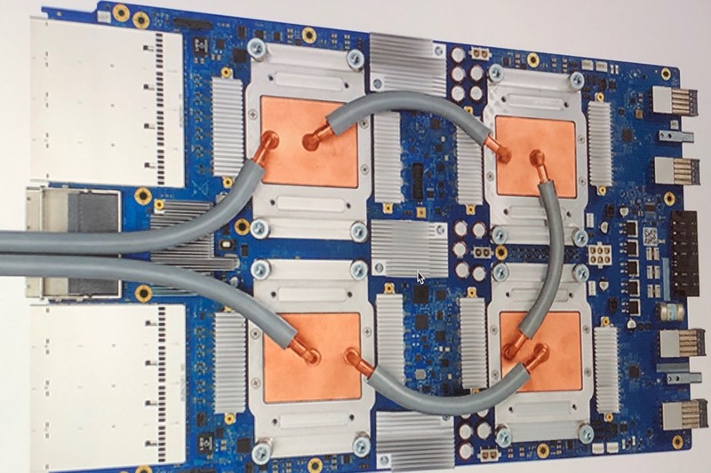 Close-up of a blue printed circuit board with orange thermal pads and gray liquid cooling tubes, representing advanced AI accelerator hardware