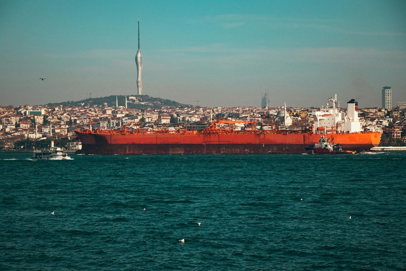 A large red oil tanker navigates through the Bosphorus Strait with Istanbul's skyline and Çamlıca Tower in the background, flanked by smaller escort vessels.