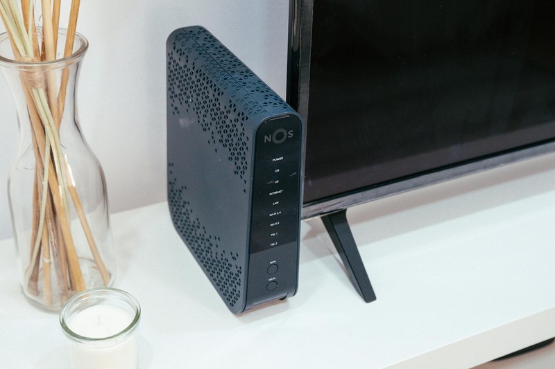 A black home router sits on a white shelf beneath a flat-screen TV in a modern living room