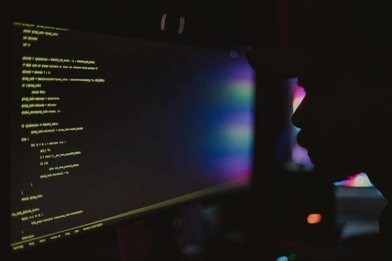 Silhouette of a programmer coding at a computer screen in a dark room with colorful ambient lighting