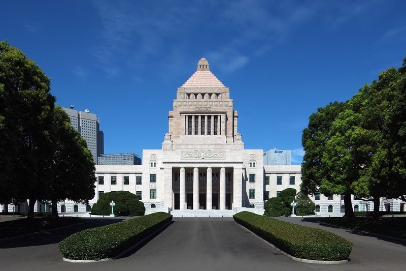 The National Diet Building in Tokyo, Japan, with its distinctive central tower and symmetrical neoclassical facade, viewed from the front approach under a clear blue sky.