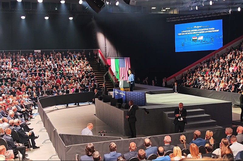 A speaker addresses a large crowd at the Day of Hungarian-American Friendship event in Budapest, with Hungarian and American flags on stage