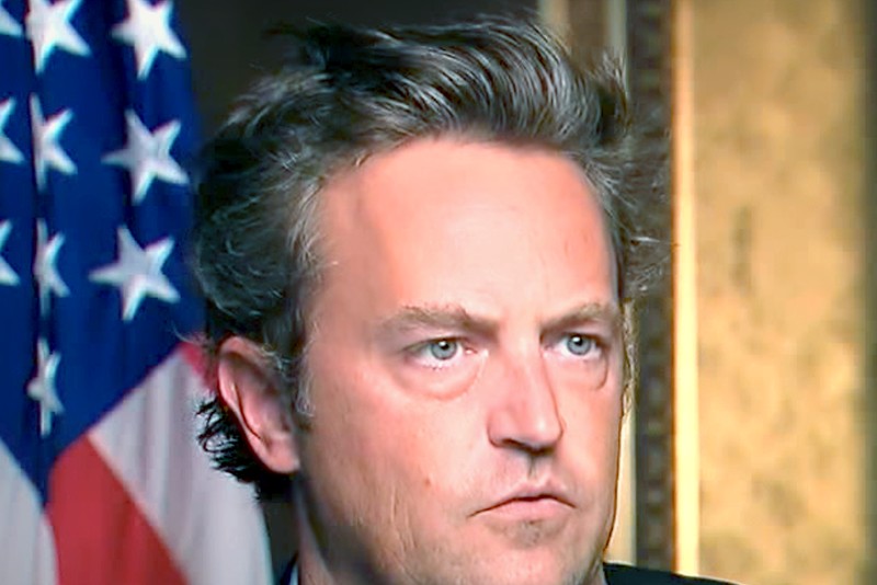 Matthew Perry speaking in a formal setting with an American flag in the background