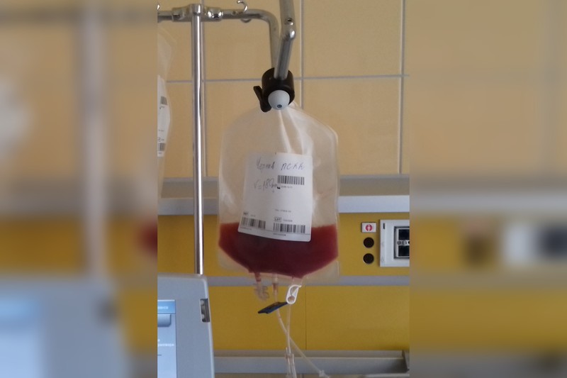 A transparent IV fluid bag hanging from a metal pole with a roller clamp in a tiled clinical setting