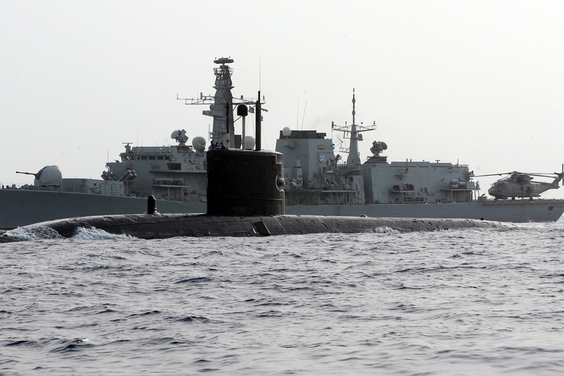 A military submarine underway on the ocean surface with a warship visible in the background under overcast skies