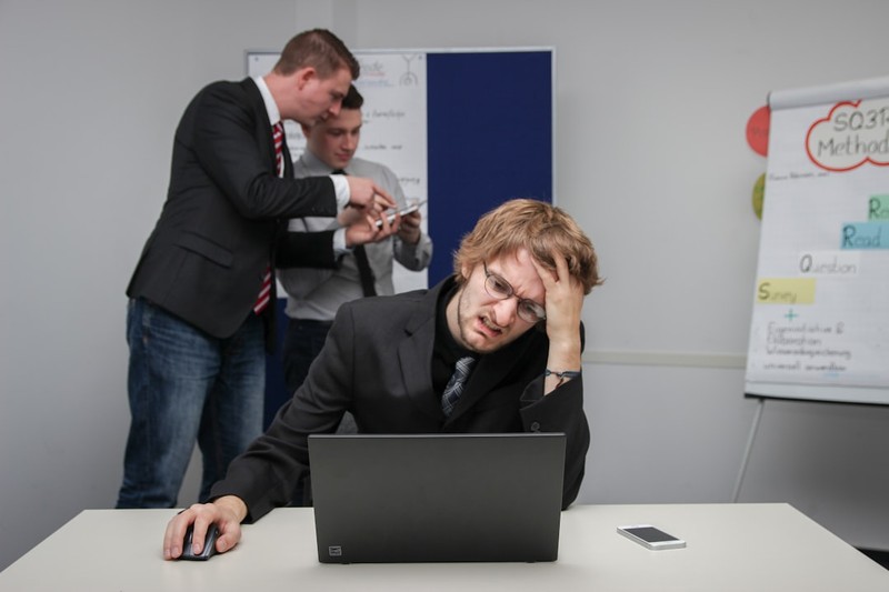 A stressed office worker sits at a desk with hand on forehead while colleagues confer in the background.