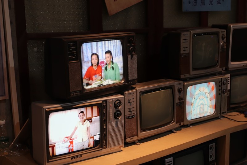 Multiple vintage CRT televisions arranged on a wooden shelf, some displaying broadcast content