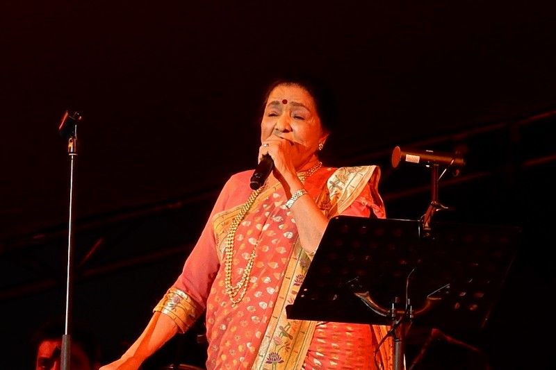 Asha Bhosle performing on stage in a red saree, singing into a microphone