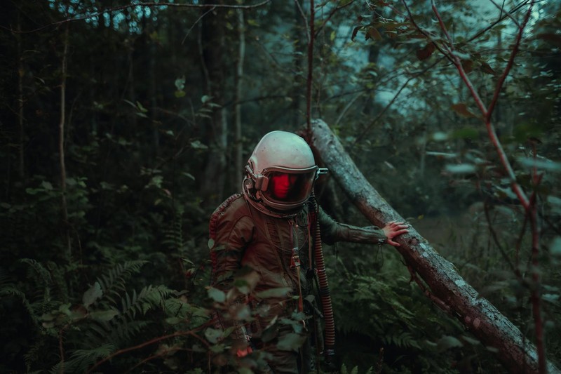 An astronaut in a retro spacesuit with a glowing red visor stands alone in a dark, mysterious forest