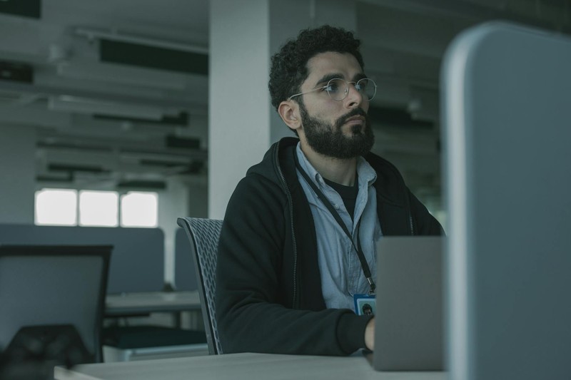 A bearded man in glasses works at a computer desk in a modern office, gazing thoughtfully at his screen