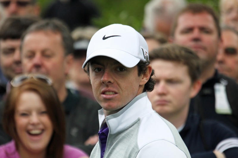 Rory McIlroy wearing a white Nike cap and jacket at a golf tournament, with a crowd of onlookers behind him