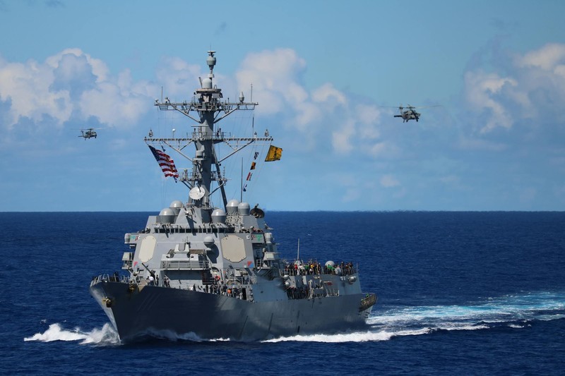 A US Navy destroyer sails on the open ocean with military helicopters flying overhead