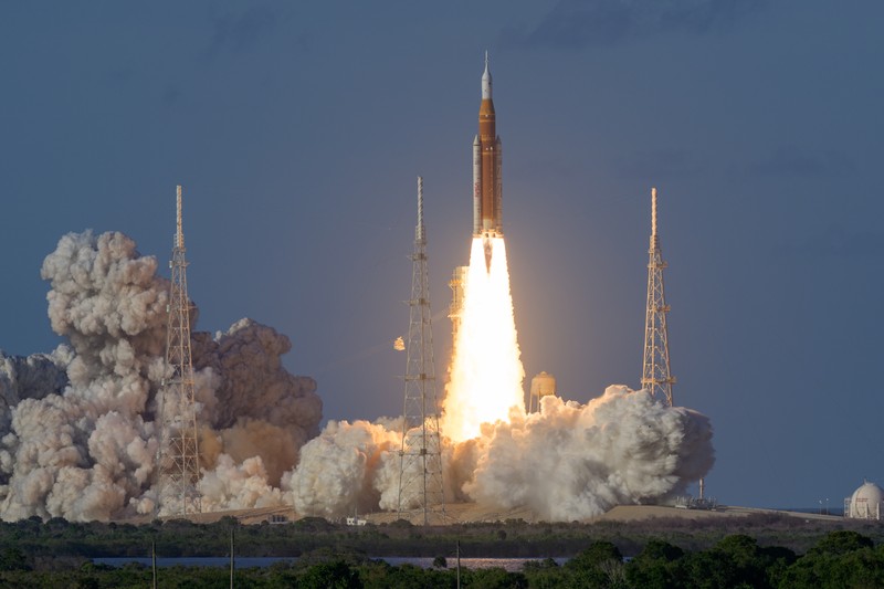NASA's Space Launch System rocket launches carrying the Orion spacecraft on the Artemis II mission, ascending amid flames and smoke against a twilight sky.