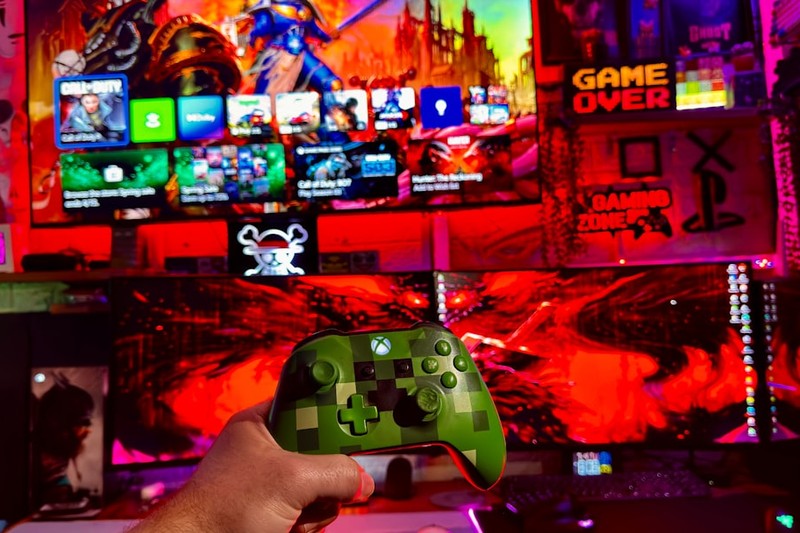 Hands holding an Xbox controller in front of a multi-screen gaming setup with neon lighting