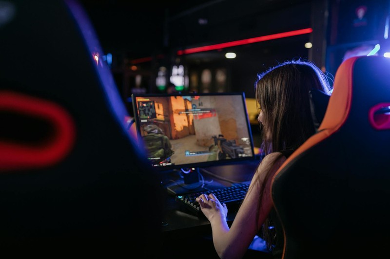 A woman deeply focused on playing a PC game in a dimly lit gaming setup with ambient lighting