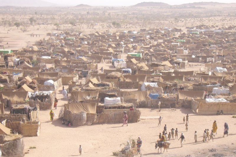 A sprawling refugee camp in an arid desert landscape with densely packed makeshift shelters and scattered residents