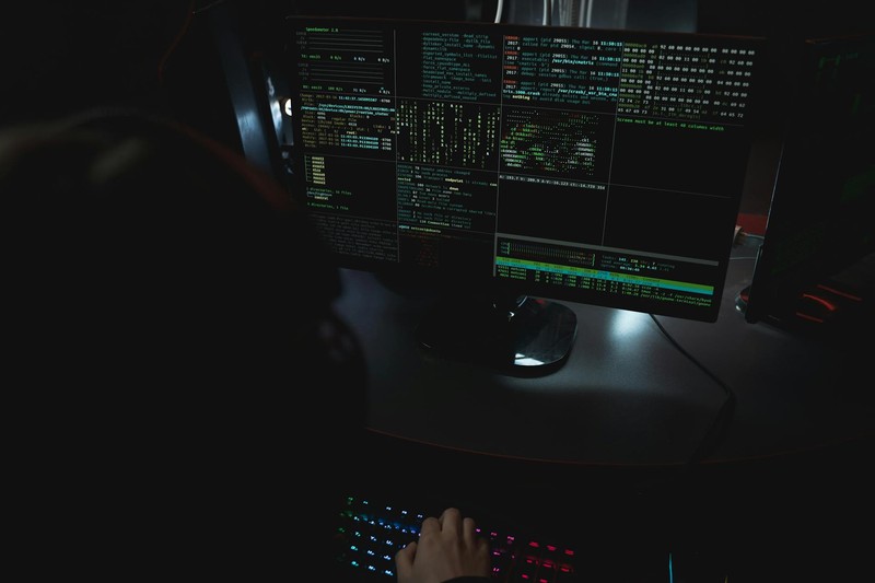 A dimly lit cybersecurity workspace with multiple monitors displaying code and terminal output, a backlit keyboard, and a person typing in a dark hoodie