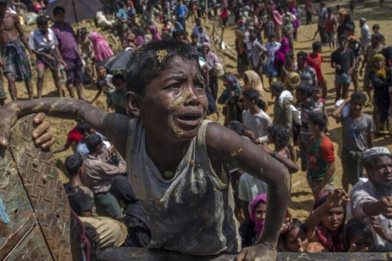 A young Rohingya boy, visibly distressed and covered in dirt, stands before a crowded camp in Cox's Bazar, Bangladesh.