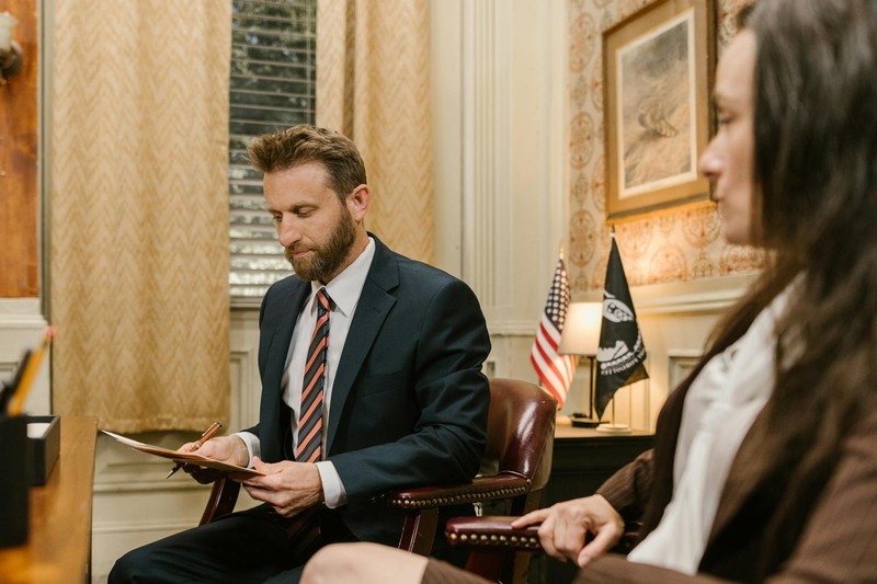 A man in a suit reviews documents in a formal office with an American flag visible