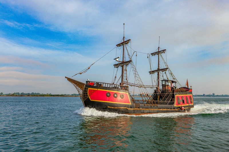 A vibrant multi-masted pirate ship sailing across blue-green waters under a partly cloudy sky