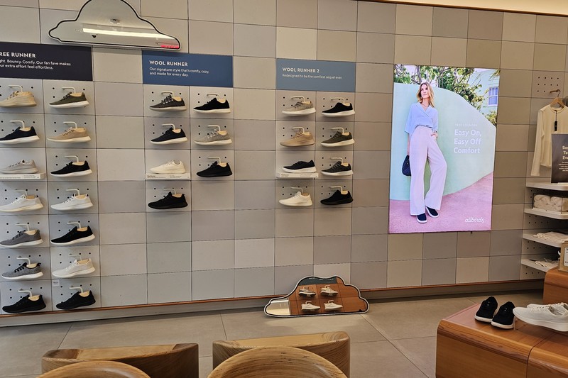 Interior of an Allbirds retail store showing shoe displays on wall-mounted shelves and a promotional screen