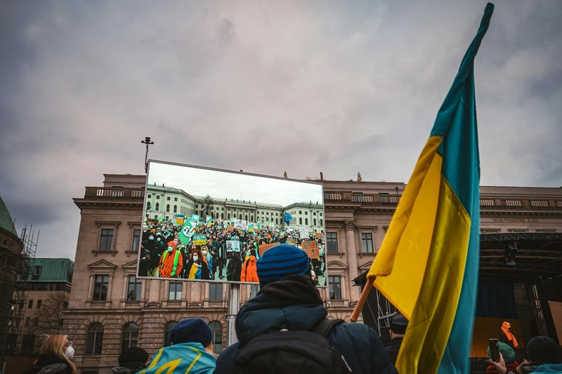 Crowd gathered at a solidarity rally in Berlin, with a Ukrainian flag held aloft against the backdrop of a classical building with a large screen.