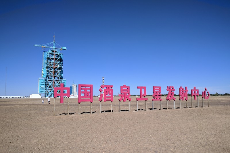 Sign for the China Jiuquan Satellite Launch Center with a launch tower in the background under a clear blue sky