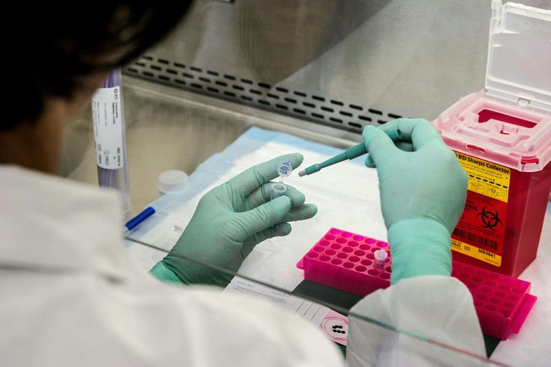 A scientist in a lab coat and green gloves handles small vials and pipettes at a biosafety cabinet in a sterile laboratory.