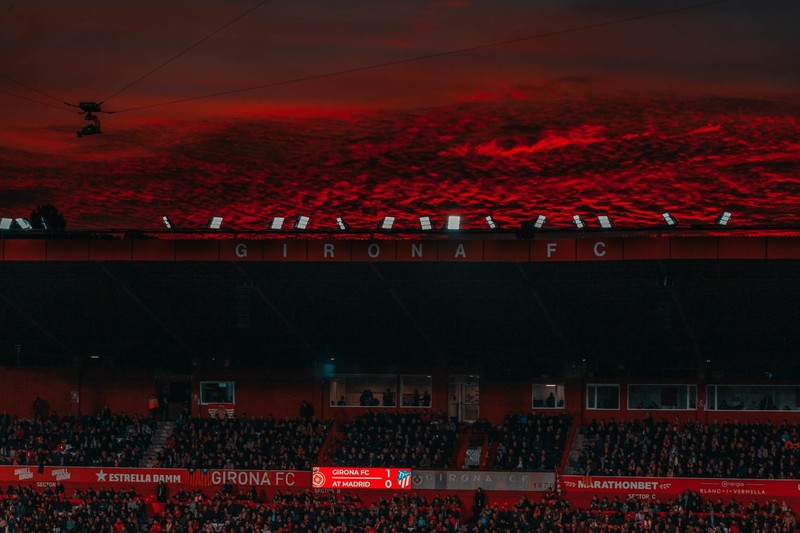 A football stadium bathed in dramatic red lighting under a sunset sky, with fans in the stands