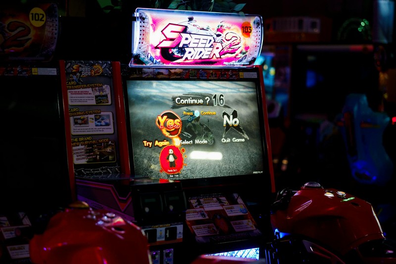 An illuminated arcade racing game machine in a dimly lit amusement center with neon pink and blue signage