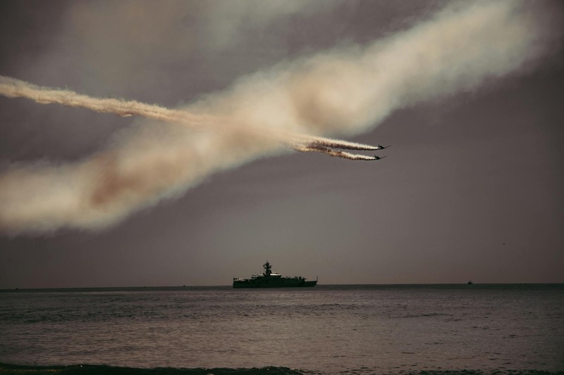 A military jet trails smoke over a naval warship on the open ocean under a dark overcast sky