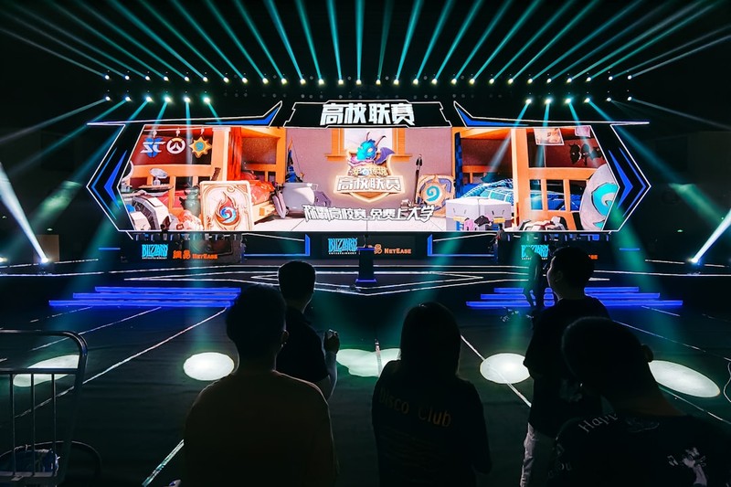 A large esports event stage with an illuminated LED screen displaying game graphics, watched by a silhouetted audience