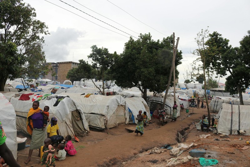 An overview of an African refugee camp with makeshift tents, families, and children gathered along dirt paths under overcast skies
