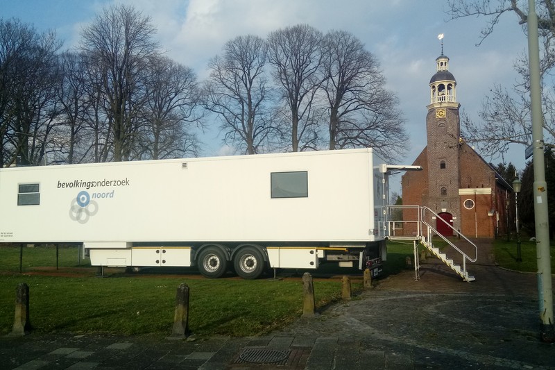 A white mobile cancer screening trailer with Dutch signage parked near a brick church in a Dutch village