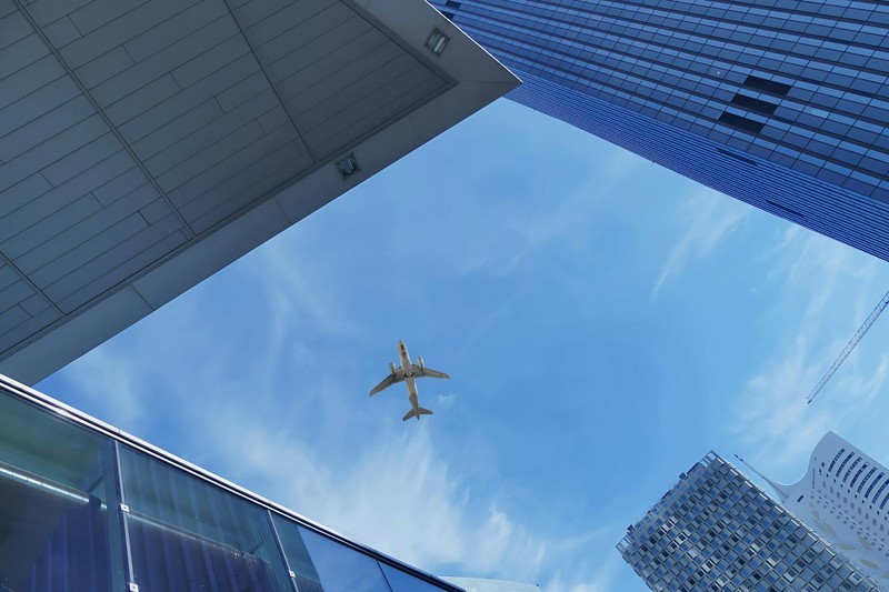 A commercial airplane flies over modern skyscrapers against a bright blue sky