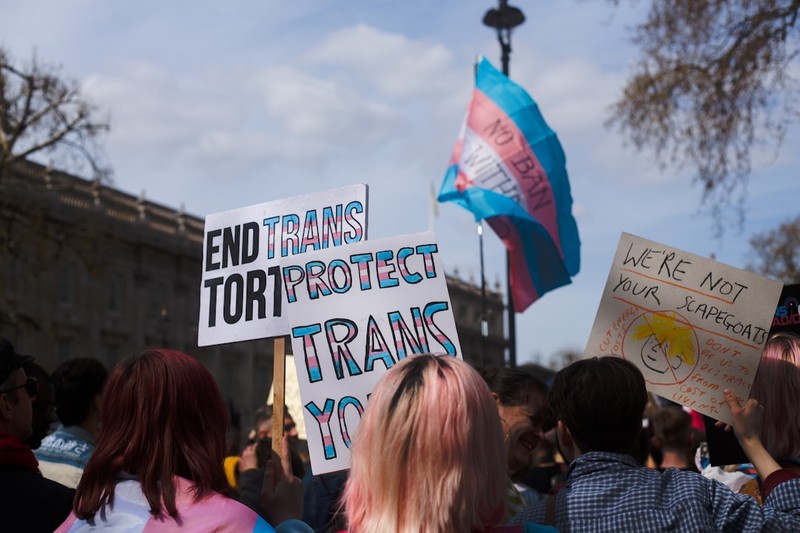 Protesters at a trans rights rally holding signs reading 'Protect Trans Youth' and 'End Trans Torture' with a transgender pride flag