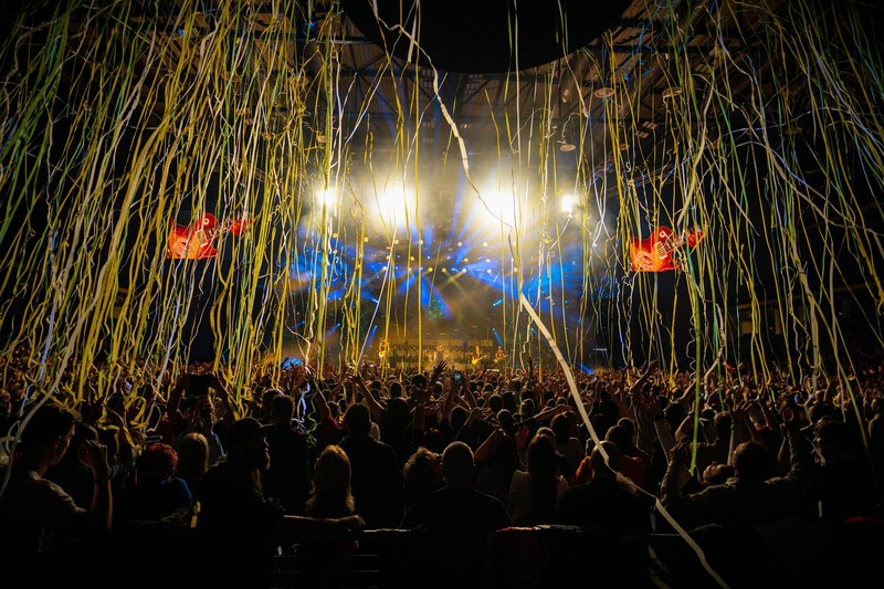 A packed concert crowd watches a vibrant stage performance with cascading confetti streamers and dramatic lighting
