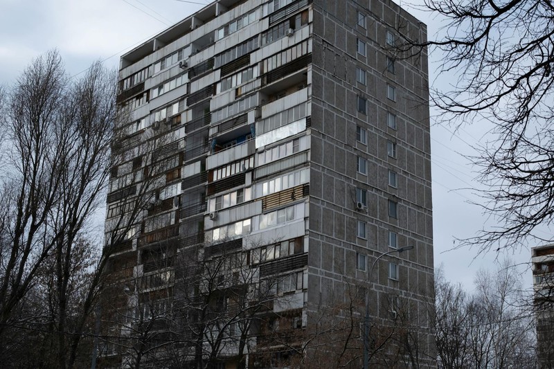 A weathered post-Soviet panelka high-rise building under a gray overcast sky, framed by bare trees