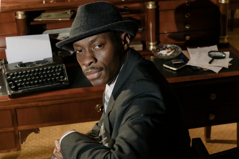 A noir detective in a fedora sits at a vintage wooden desk with a typewriter and magnifying glass in a dimly lit office