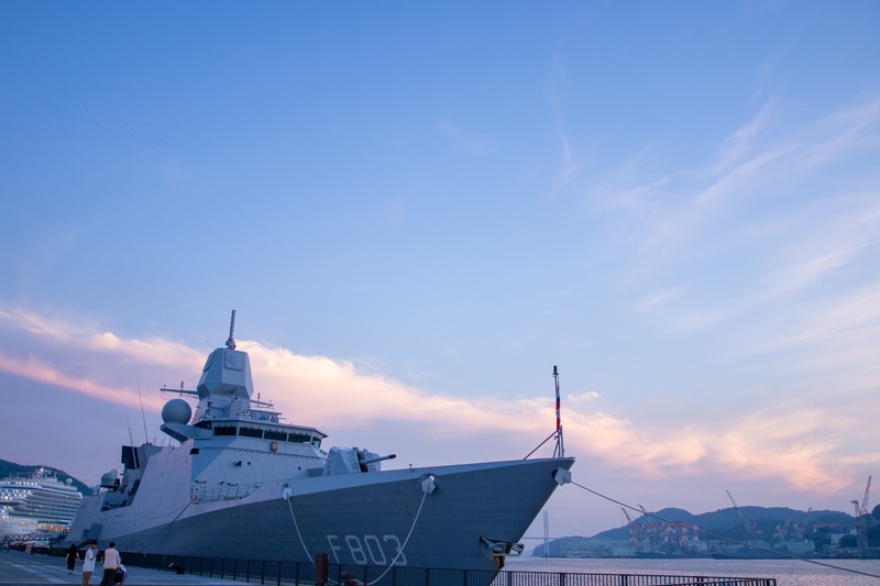 A naval warship docked at the Nagasaki Shipyard of Mitsubishi Heavy Industries at twilight, with industrial port infrastructure and hills visible in the background.