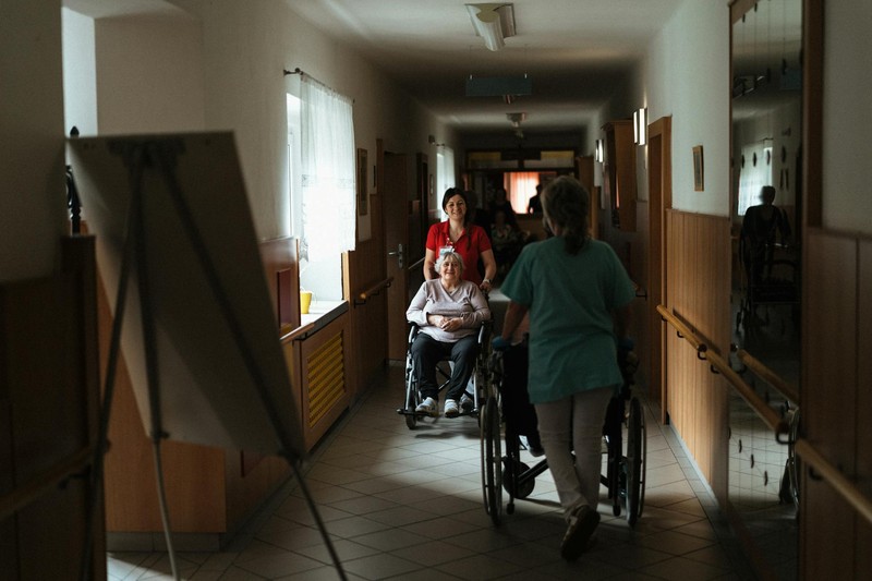 A caregiver pushes an elderly woman in a wheelchair down a nursing home corridor while another caregiver assists a second resident