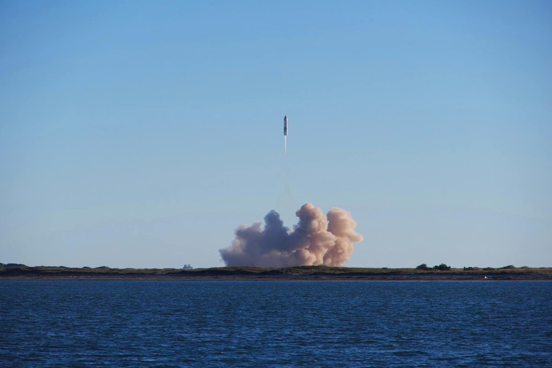 A rocket launches over coastal waters, leaving a massive smoke plume against a clear blue sky