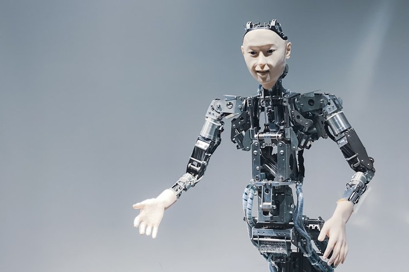 A humanoid robot with exposed mechanical components and a human-like face stands in a studio setting