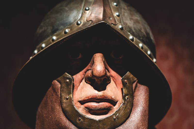 A weathered medieval metal helmet with riveted details, lit with dramatic shadows against a dark reddish-brown background